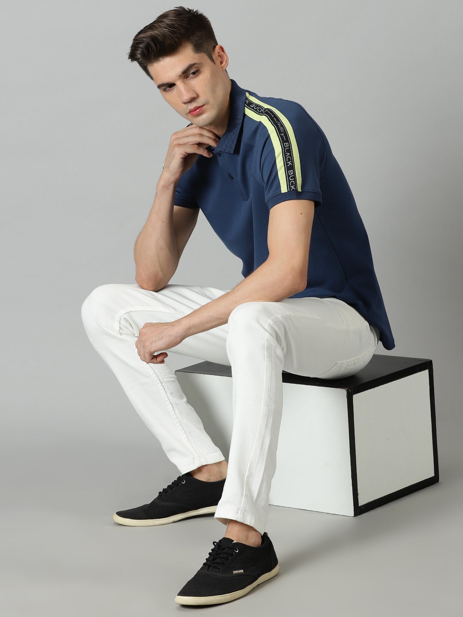 Man sitting on a modern chair wearing a navy polo shirt with yellow stripes, white pants, and black shoes.
